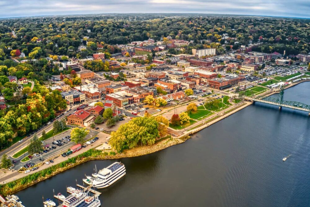 Aerial view of Stillwater, Oklahoma, highlighting the scenic downtown area near the waterfront, lush greenery, historic buildings, and boats docked along the river, perfect for tourists exploring Payne County.