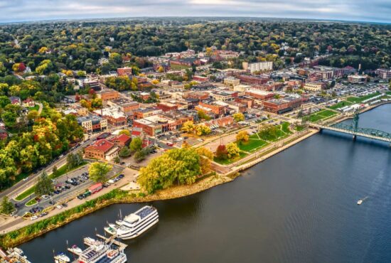 Aerial view of Stillwater, Oklahoma, highlighting the scenic downtown area near the waterfront, lush greenery, historic buildings, and boats docked along the river, perfect for tourists exploring Payne County.